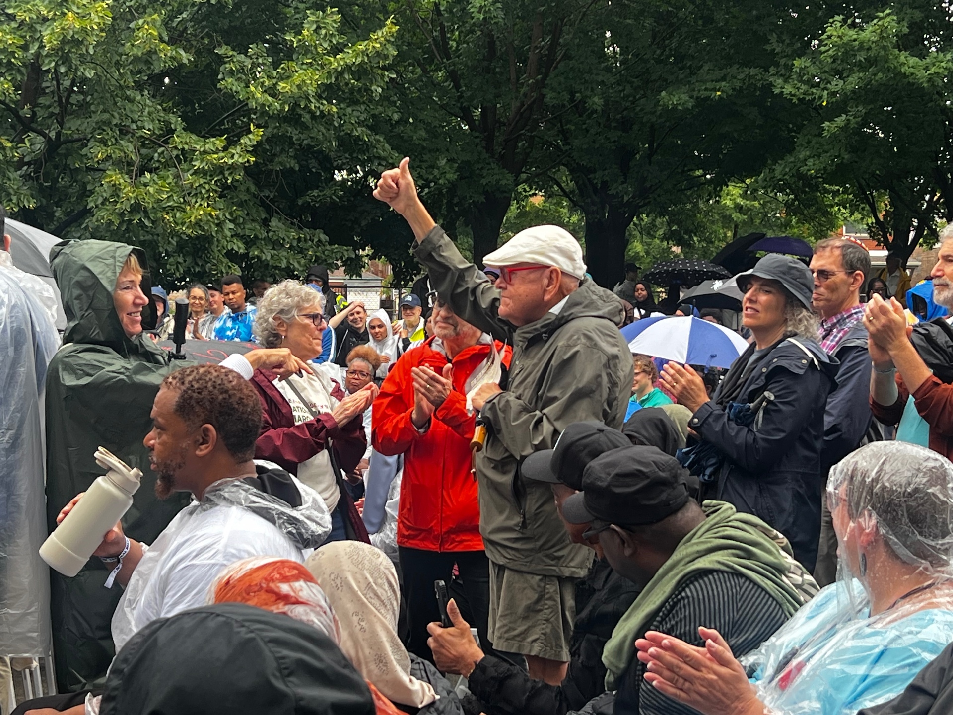 Rain-soaked crowd in ponchos applauding speakers under the trees at Marquette Park
